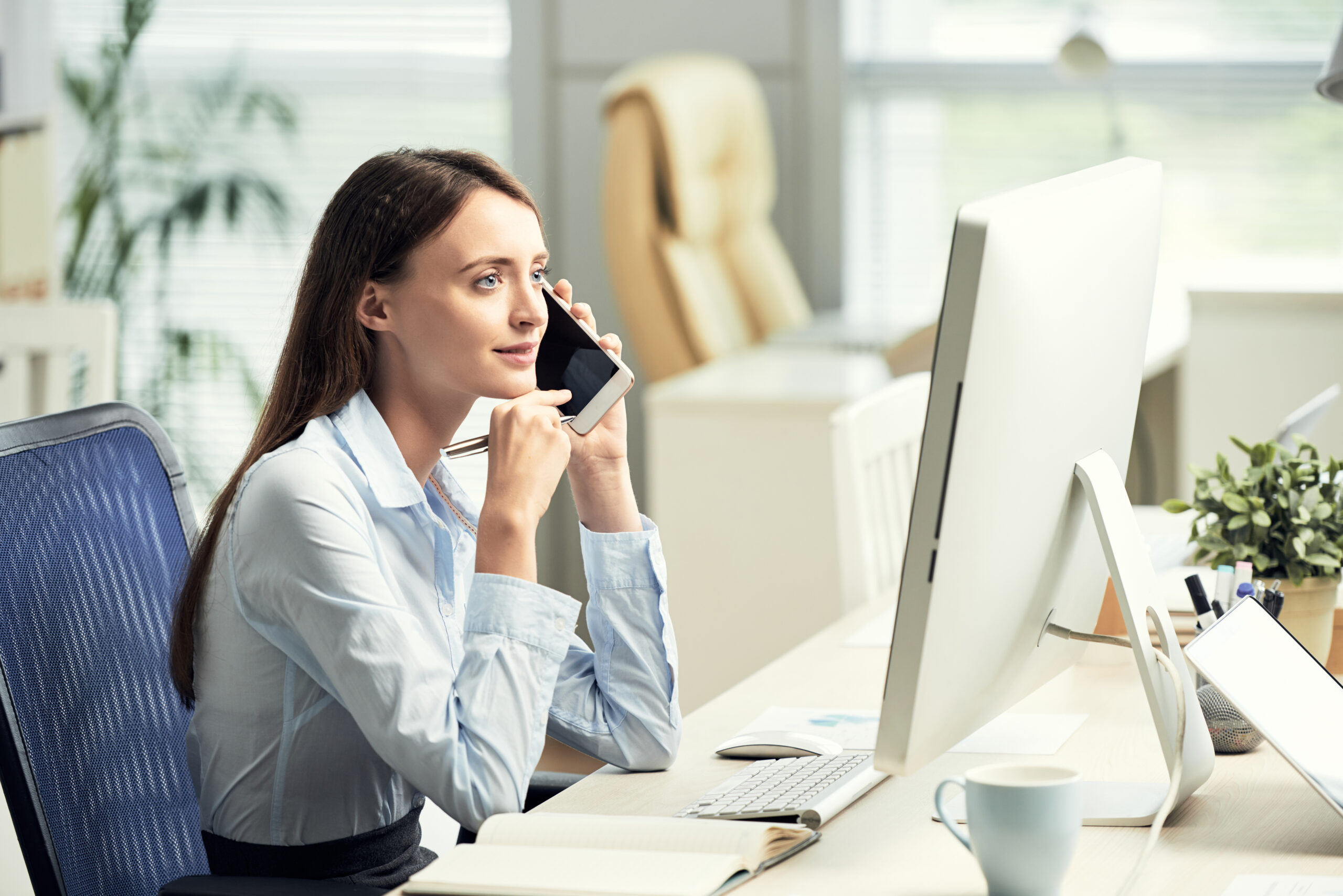 caucasian-female-office-worker-sitting-desk-front-screen-talking-smartphone